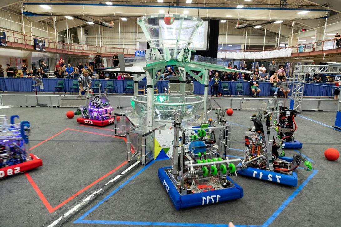 Four FIRST Robotics robots complete tasks in a competition field as an audience watches